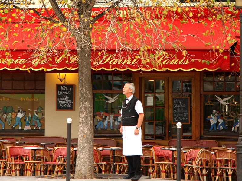 Waiter stands outside a restaurant in Paris as autumn leaves fall around him...